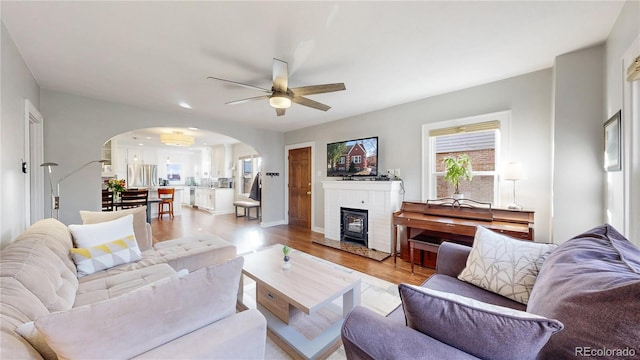 living area featuring arched walkways, baseboards, ceiling fan, light wood-style floors, and a fireplace