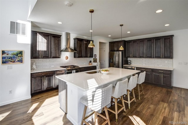 kitchen featuring sink, stainless steel appliances, wall chimney exhaust hood, pendant lighting, and a kitchen island with sink