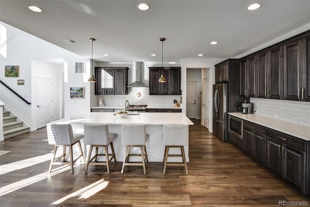 kitchen with an island with sink, stainless steel appliances, wall chimney range hood, and hanging light fixtures