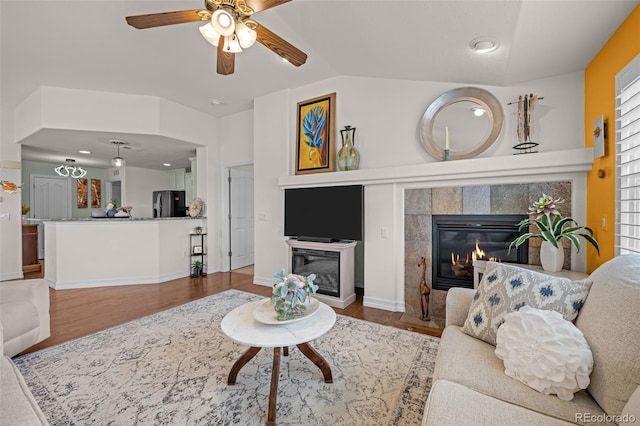 living room with baseboards, vaulted ceiling, a tile fireplace, wood finished floors, and a ceiling fan