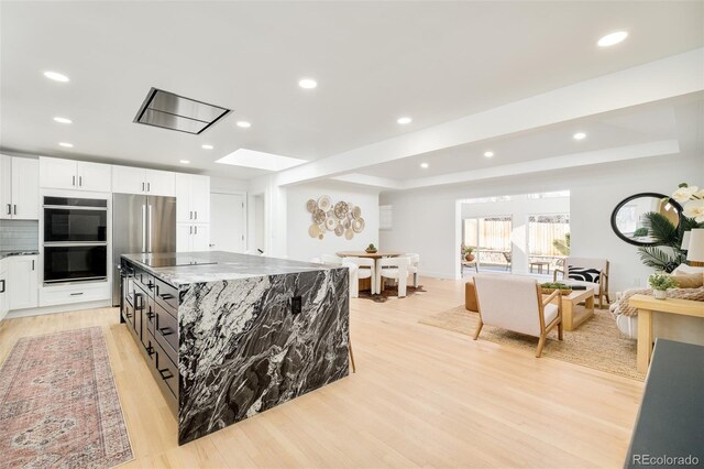 kitchen with dobule oven black, a kitchen island, white cabinetry, dark stone counters, and light wood-style flooring
