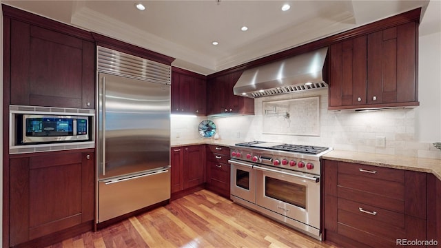 kitchen with built in appliances, wall chimney exhaust hood, decorative backsplash, light hardwood / wood-style floors, and light stone counters