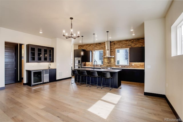 kitchen featuring wine cooler, a breakfast bar, light wood finished floors, a notable chandelier, and wall chimney exhaust hood