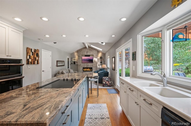 kitchen featuring black appliances, a stone fireplace, a sink, lofted ceiling, and light wood-style floors