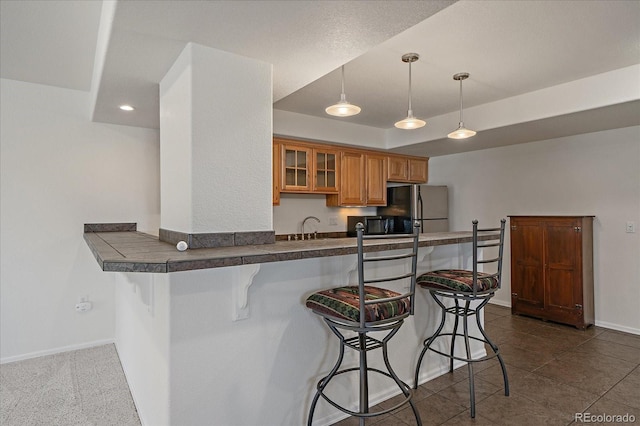 kitchen featuring tile countertops, glass insert cabinets, freestanding refrigerator, a peninsula, and a kitchen bar