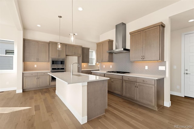 kitchen featuring appliances with stainless steel finishes, wall chimney exhaust hood, sink, decorative light fixtures, and a center island with sink