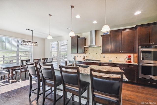 kitchen featuring pendant lighting, backsplash, a kitchen island with sink, stainless steel appliances, and wall chimney exhaust hood