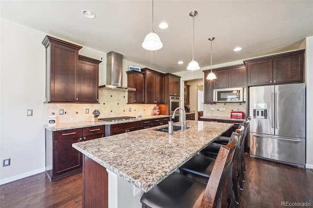 kitchen with sink, appliances with stainless steel finishes, hanging light fixtures, an island with sink, and wall chimney exhaust hood