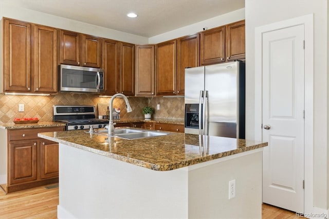 kitchen featuring sink, dark stone counters, a center island with sink, appliances with stainless steel finishes, and light wood-type flooring