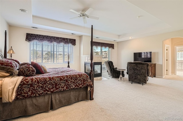 carpeted bedroom with visible vents, a ceiling fan, a tray ceiling, arched walkways, and a tile fireplace