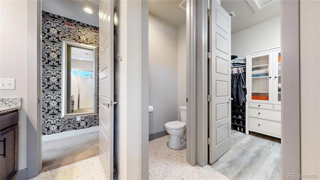 bathroom with a walk in closet, toilet, vanity, baseboards, and wallpapered walls