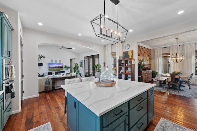 kitchen with ceiling fan with notable chandelier, a kitchen island, hanging light fixtures, dark wood-type flooring, and light stone counters