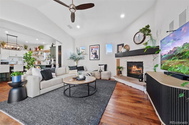 living room featuring lofted ceiling, hardwood / wood-style floors, a stone fireplace, and ceiling fan with notable chandelier