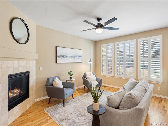 living area with light wood finished floors, a tile fireplace, a ceiling fan, and baseboards