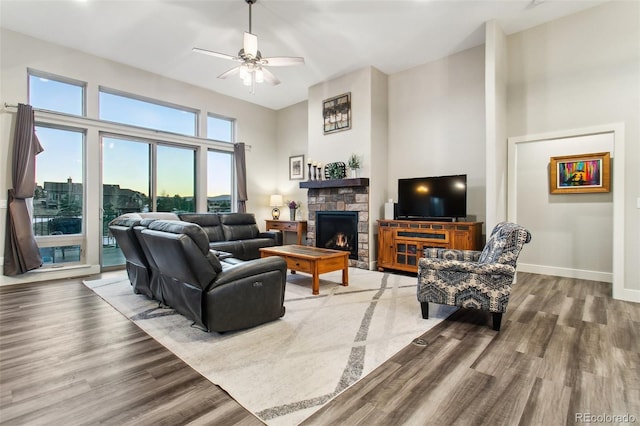 living room featuring wood finished floors, baseboards, ceiling fan, a stone fireplace, and a towering ceiling