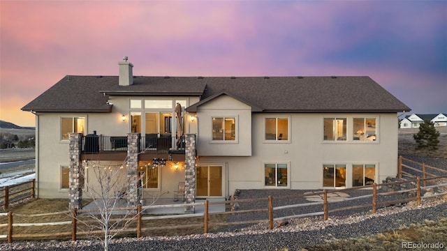 back of house at dusk with fence, a shingled roof, stucco siding, a chimney, and a patio area