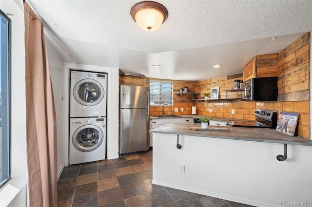 kitchen with a sink, a textured ceiling, stainless steel appliances, a peninsula, and stacked washer / dryer