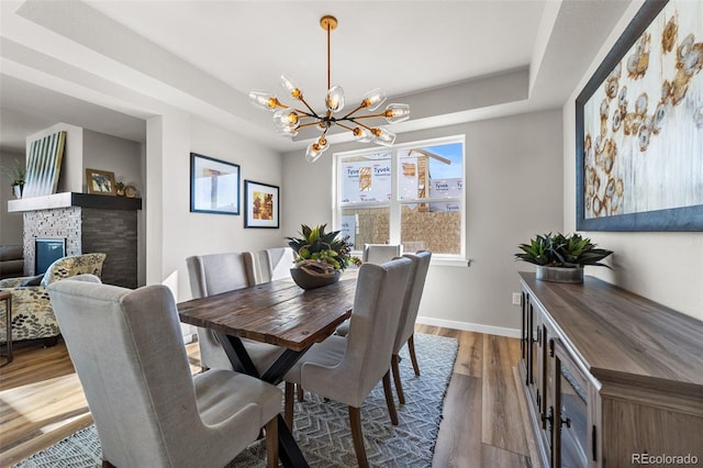 dining area featuring a raised ceiling, wood finished floors, a stone fireplace, an inviting chandelier, and baseboards