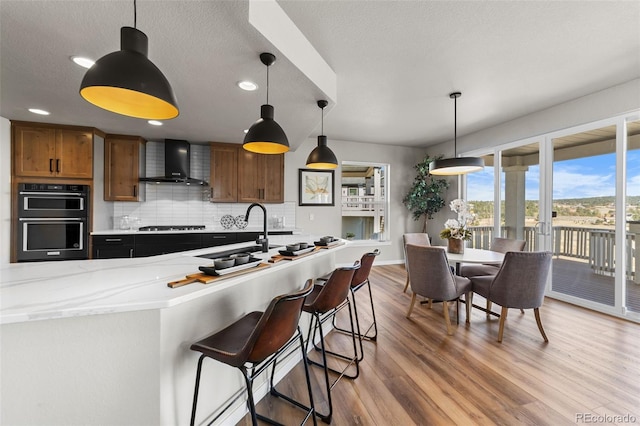 kitchen featuring tasteful backsplash, a sink, dobule oven black, wall chimney exhaust hood, and light wood-type flooring