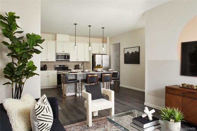kitchen featuring stainless steel appliances, a center island with sink, tasteful backsplash, dark wood-style flooring, and baseboards