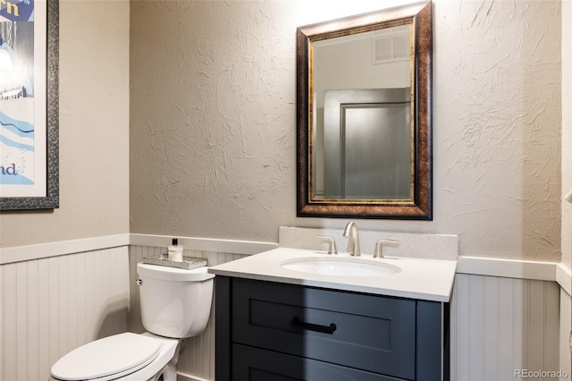 bathroom with vanity, visible vents, wainscoting, toilet, and a textured wall