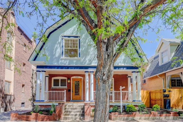 view of front of house with covered porch, a gambrel roof, brick siding, and fence