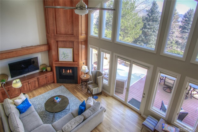 living room featuring a towering ceiling, ceiling fan, a glass covered fireplace, and light wood-style flooring