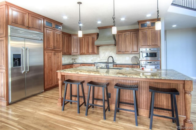 kitchen featuring stainless steel appliances, custom range hood, a center island with sink, and light stone countertops