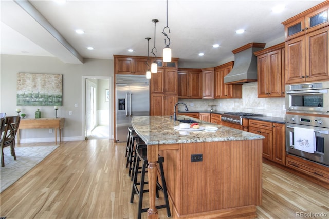 kitchen with dark stone counters, an island with sink, appliances with stainless steel finishes, wall chimney range hood, and a sink