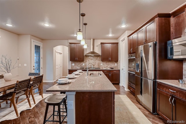 kitchen featuring tasteful backsplash, arched walkways, wall chimney exhaust hood, appliances with stainless steel finishes, and wood finished floors