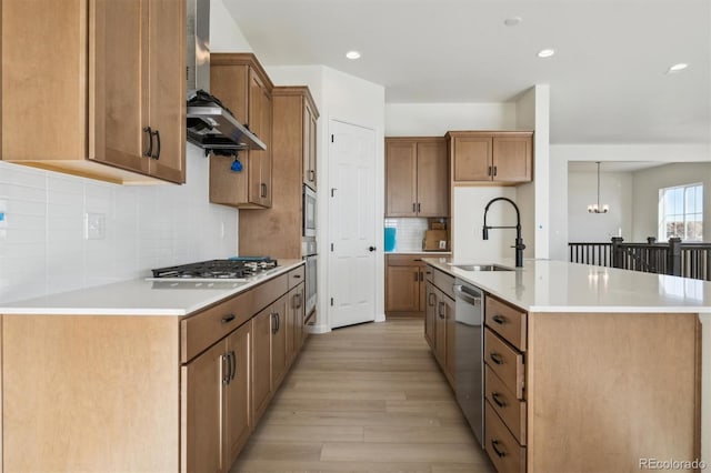 kitchen featuring sink, wall chimney exhaust hood, stainless steel appliances, an inviting chandelier, and an island with sink