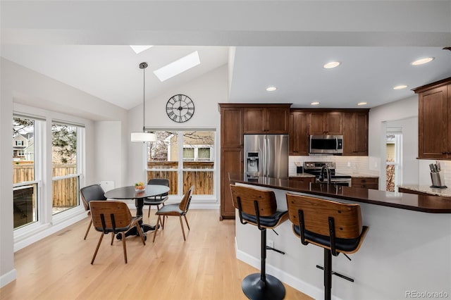 kitchen with light wood finished floors, tasteful backsplash, hanging light fixtures, appliances with stainless steel finishes, and vaulted ceiling with skylight