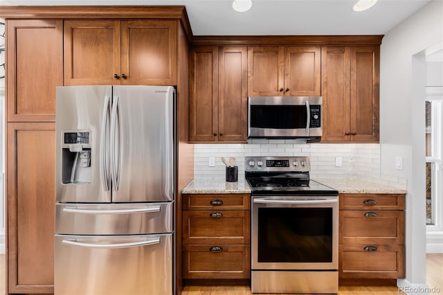 kitchen featuring stainless steel appliances, brown cabinetry, backsplash, and light stone countertops