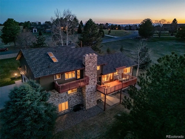 back of property at dusk with a shingled roof, stone siding, a balcony, and a chimney
