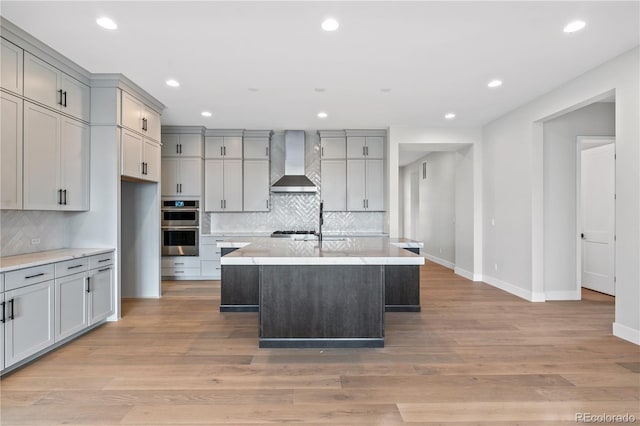 kitchen featuring gray cabinets, double oven, an island with sink, light hardwood / wood-style floors, and wall chimney exhaust hood