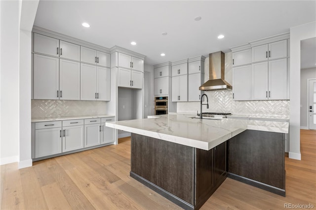 kitchen featuring sink, light stone counters, light wood-type flooring, an island with sink, and wall chimney range hood