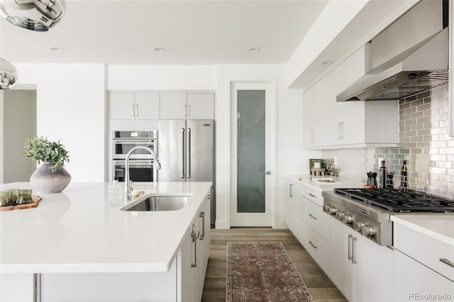 kitchen with backsplash, wall chimney range hood, sink, an island with sink, and stainless steel appliances