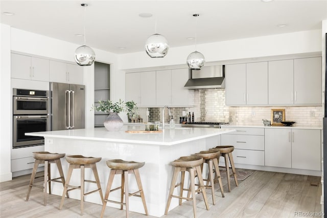 kitchen with pendant lighting, a center island with sink, wall chimney exhaust hood, white cabinetry, and stainless steel appliances