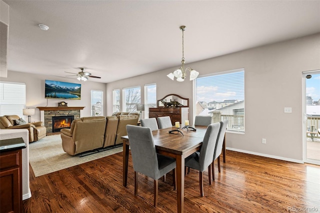 dining room with a stone fireplace, wood-type flooring, and ceiling fan with notable chandelier