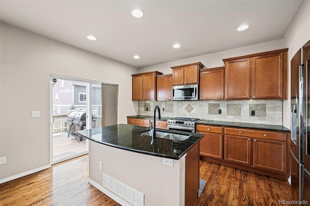 kitchen with sink, a center island with sink, appliances with stainless steel finishes, dark hardwood / wood-style floors, and dark stone counters