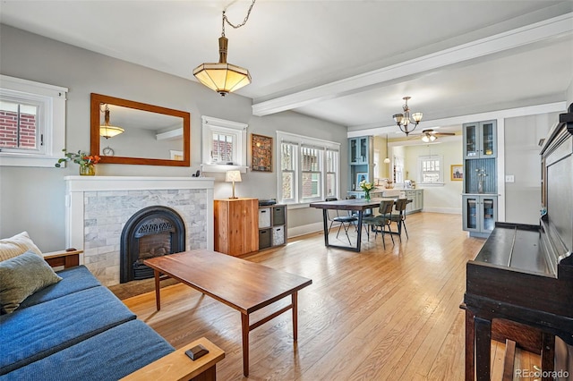 living area featuring beam ceiling, baseboards, light wood-style flooring, a notable chandelier, and a glass covered fireplace