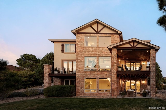 back house at dusk with a balcony, a patio area, and a lawn