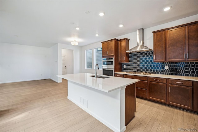 kitchen featuring sink, wall chimney exhaust hood, light wood-type flooring, an island with sink, and appliances with stainless steel finishes