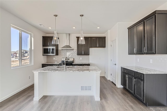 kitchen featuring appliances with stainless steel finishes, decorative light fixtures, an island with sink, light stone countertops, and wall chimney exhaust hood