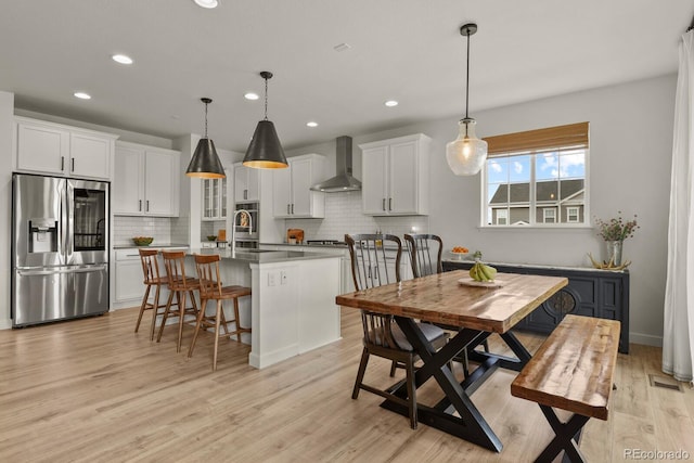 kitchen featuring white cabinetry, hanging light fixtures, wall chimney range hood, stainless steel refrigerator with ice dispenser, and a center island with sink