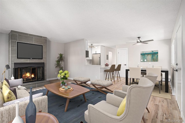 living room with ceiling fan, a tiled fireplace, a textured ceiling, and light hardwood / wood-style flooring