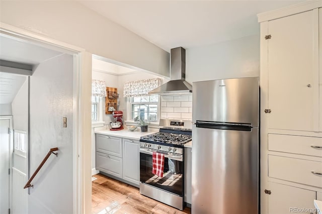 kitchen with decorative backsplash, light countertops, wall chimney exhaust hood, light wood finished floors, and stainless steel appliances