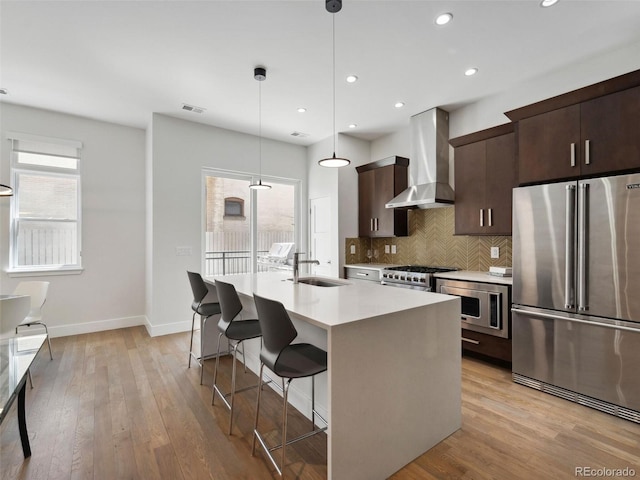 kitchen featuring sink, wall chimney range hood, decorative light fixtures, a center island with sink, and high end appliances