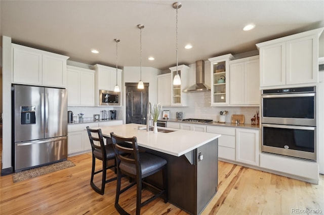 kitchen featuring appliances with stainless steel finishes, a sink, an island with sink, wall chimney exhaust hood, and white cabinetry