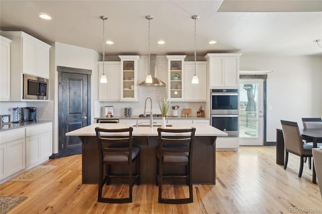 kitchen featuring light wood-style floors, a center island with sink, wall chimney range hood, light countertops, and stainless steel appliances